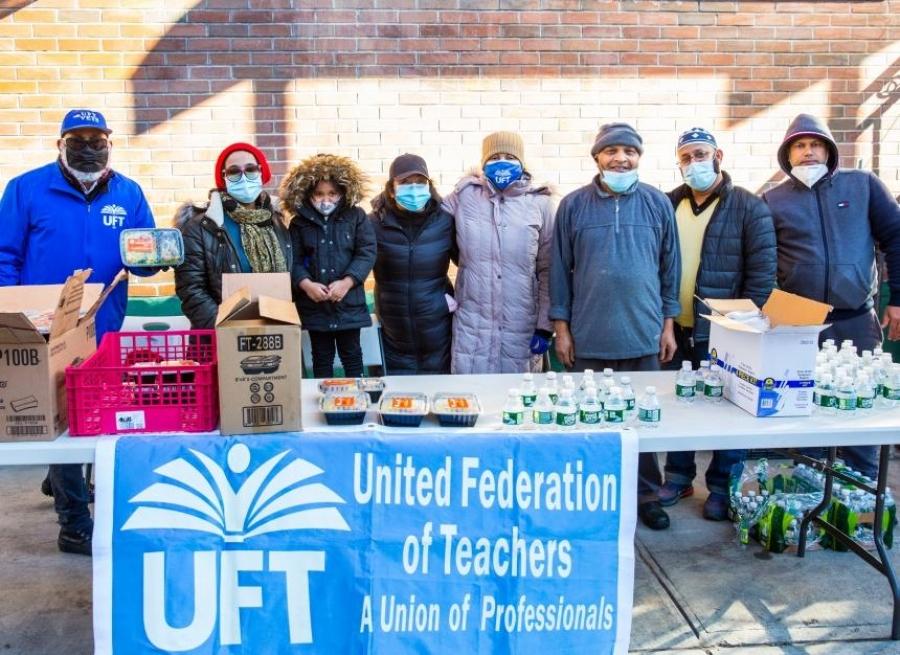 a group of adults and children wearing winter coats and face masks stand behind a table covered in packaged food with a UFT banner