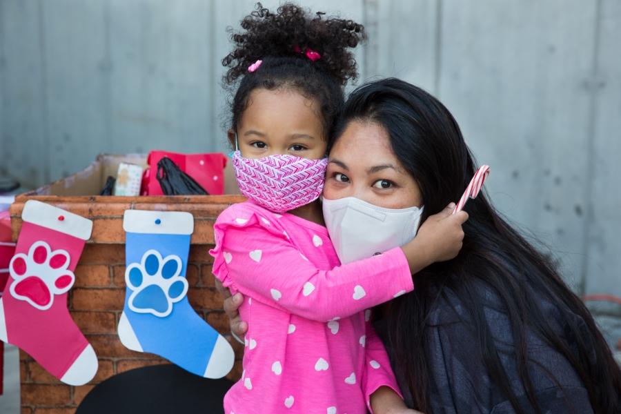 a little girl in a pink dress hugs a woman. both wear face masks. there are two holiday stockings behind them.