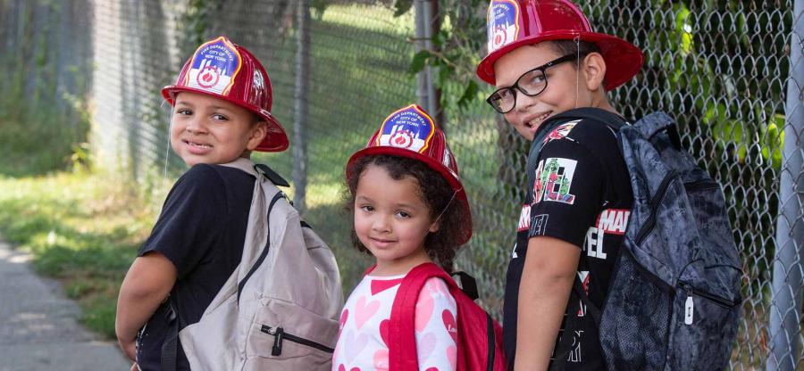 Three children wearing backpacks and fire helmets