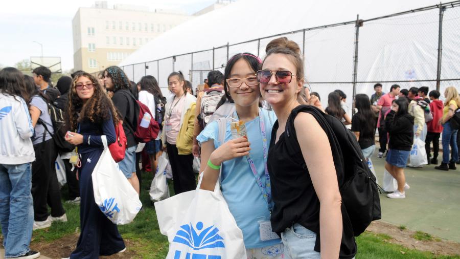 Two women with glasses pose for a photo amongst a crowd of many others in a field with a fence around it, one holding a UFT swag bag
