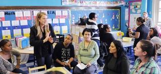 Teacher Meg O’Dwyer (standing, left) and paraprofessional Jonalyn Reyes (standing, right, back to camera) lead a professional development session for colleagues at P 352 in the Bronx. The session was set in Reyes’ classroom to explain the layout and transitioning between stations. 