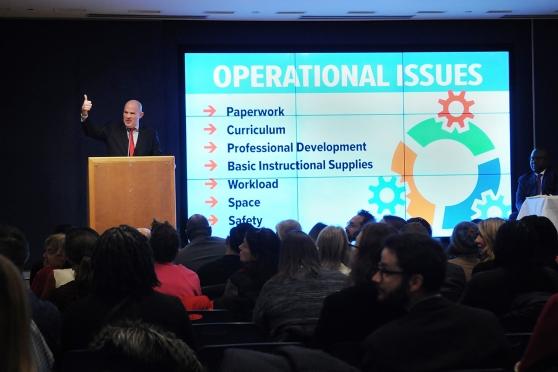 Michael Mulgrew standing in front of podium and a large display screen