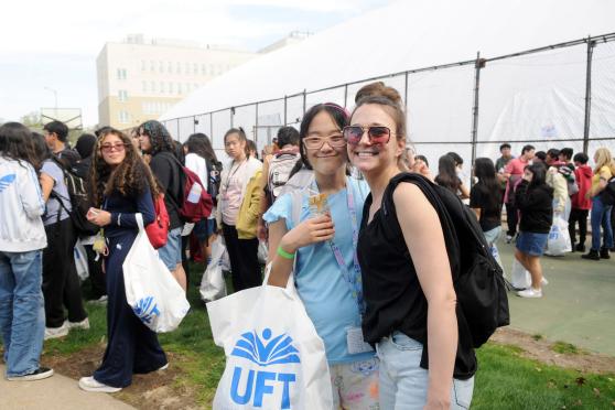 Two women with glasses pose for a photo amongst a crowd of many others in a field with a fence around it, one holding a UFT swag bag