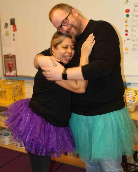 A woman wearing a purple tutu hugs a man wearing a green tutu in a classroom.