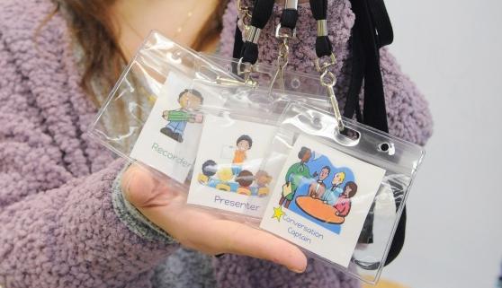A teacher holds up lanyards that remind students of their roles and tasks in small group discussions. 