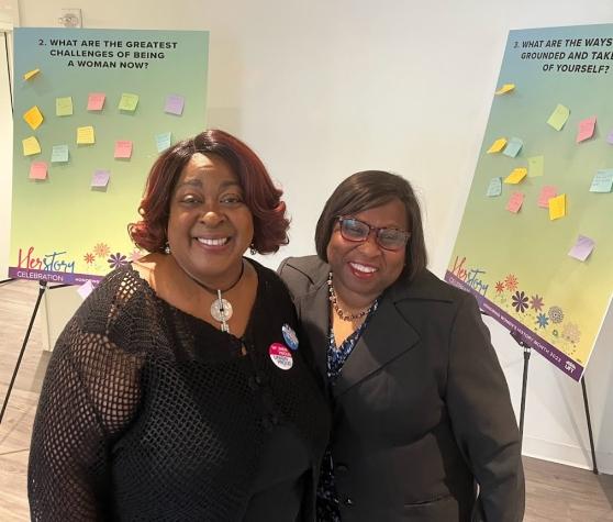Two women pose for a photo in front of a display. 