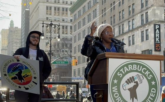 A woman stands at a podium and a man behind her is smiling and holding a sign supporting Starbucks unions