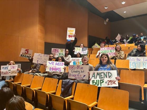Members sitting in an auditorium hold up signs demanding reallocation of charter school space. 