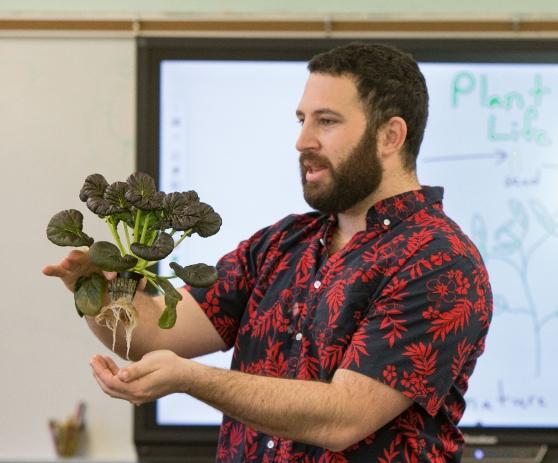 A white man with a beard wearing a flower-patterned shirt holds a leafy plant