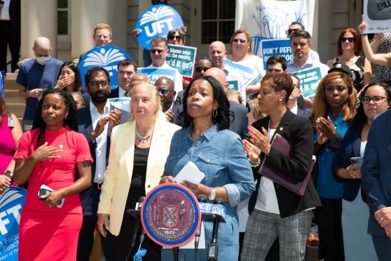 A Black woman wearing a denim dress stands at a podium with a crowd behind her