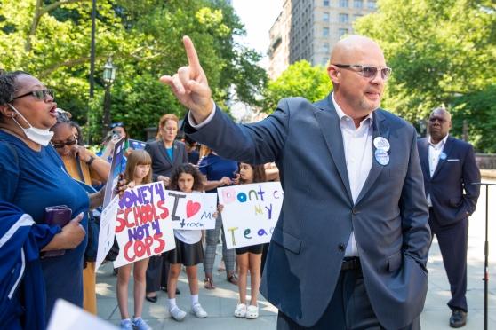 A bald man wearing a suit and glasses holds up one finger with a crowd behind him