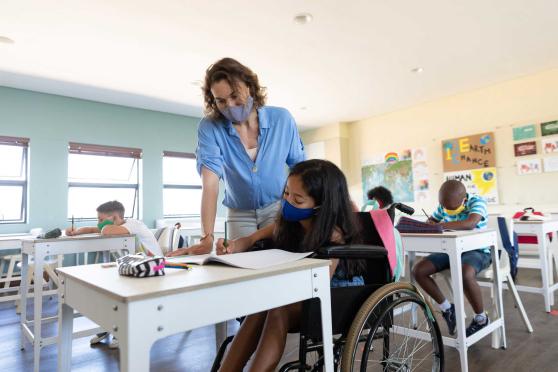 A teacher wearing a mask assists a student with writing at her desk 