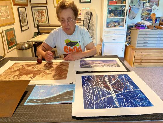 An elderly woman sits by a table in her art studio surrounded by her drawings and paintings.