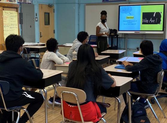 A teacher with a class of socially distanced students are engaged in a lesson. All are masked and the teacher is referencing material on the white board. 