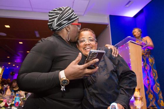 A woman on the left embraces the woman on the right with a kiss to the top of her head