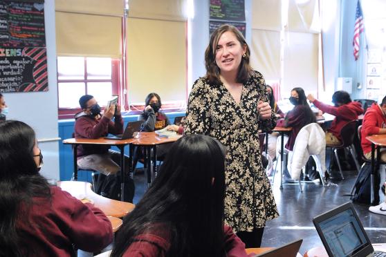 Female teacher standing in room with others who are sitting.