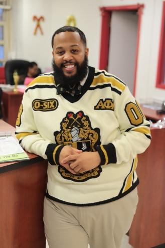 Young bearded man standing and smiling wearing a cream sweater with greek letters.